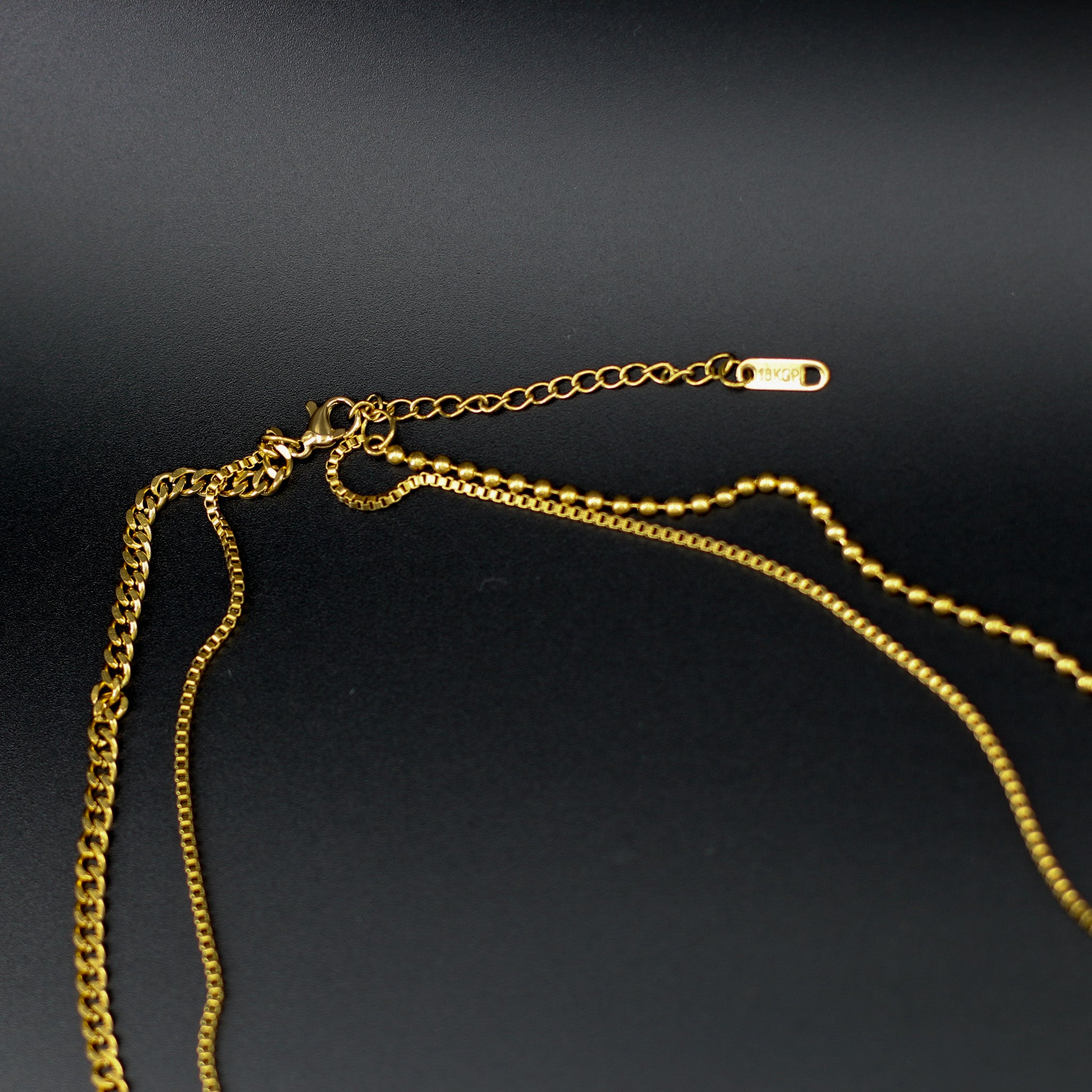 Mehrlagige Kette 'Tri-Chain Vibes' auf einem Dekolleté, mit schwarzem Stein-Anhänger, Medaillon und vergoldeten Ketten, gefertigt aus 316L Edelstahl.
