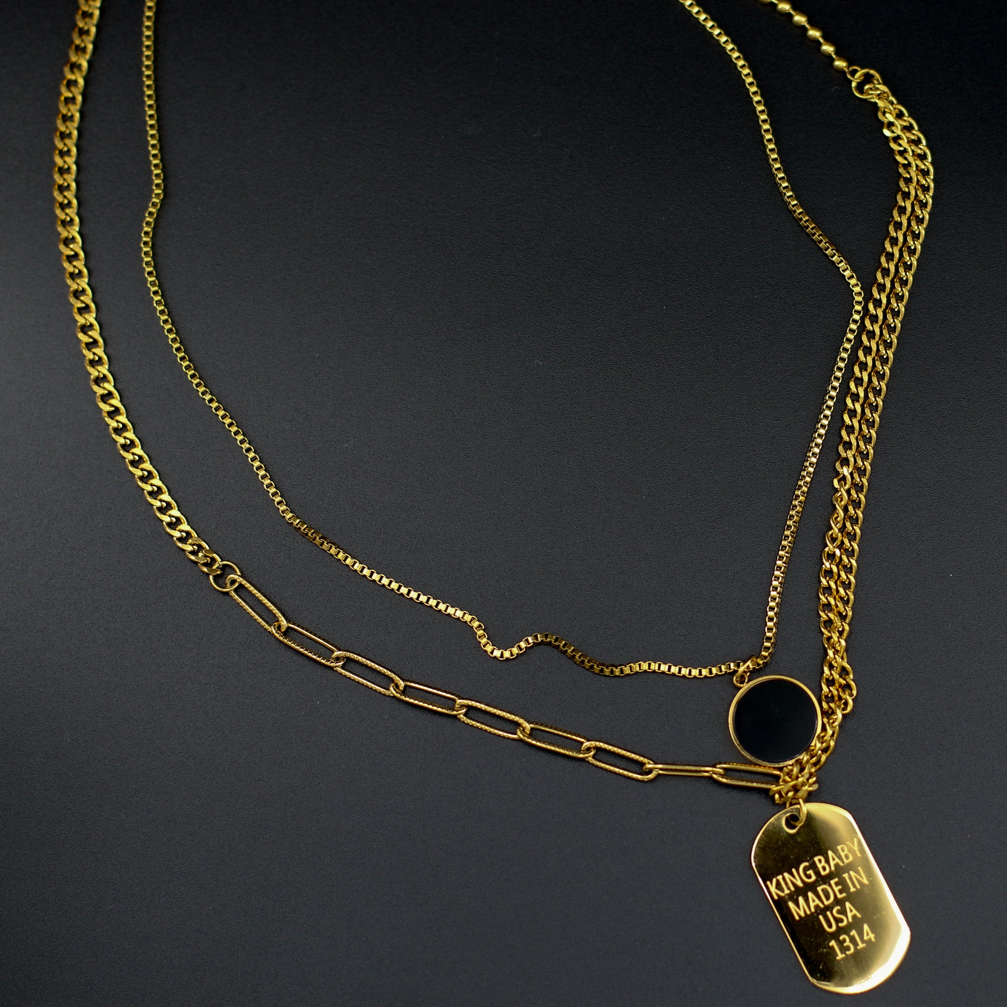 Mehrlagige Kette 'Tri-Chain Vibes' auf einem Dekolleté, mit schwarzem Stein-Anhänger, Medaillon und vergoldeten Ketten, gefertigt aus 316L Edelstahl.
